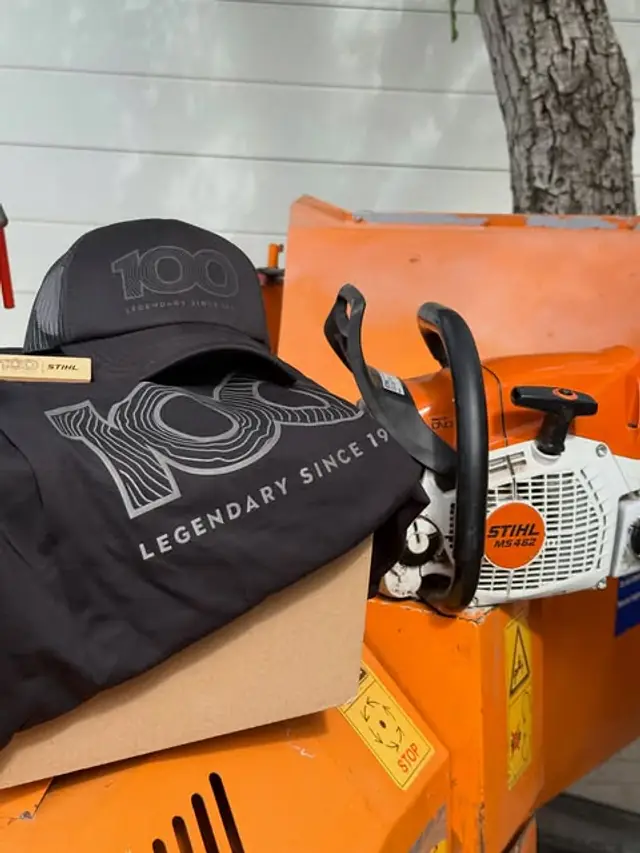 STIHL MS462 láncfűrész a narancssárga ágaprító gép tetején, mellette márkás sapka és póló.